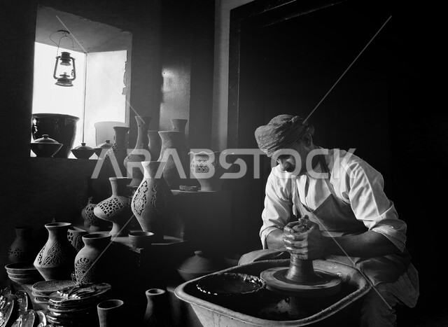 مهن وصناعات يدوية شعبية تراثية قديمة، متجر لبيع أواني الفخارات في سوق شعبي تقليدي في سلطنة عمان، الأواني والمنتجات الفخارية المصنوعة من طين، صورة باللون الأبيض والأسود لرجل عربي خليجي عماني يرتدي الدشداشه والعمامة يقوم بمهارة صنع الفخار