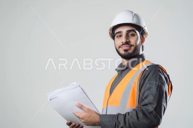 الاشراف والرقابة على المشاريع الهندسية، الاعمار بسواعد ابناء الوطن، بورتريه لمهندس عربي خليجي سعودي يرتدي الثوب التقليدي مع خوذة وسترة الوقاية يمسك مخطط البناء وينظر إليه بإيماءات التركيز، مهن ووظائف سعودية، خلفية رمادية