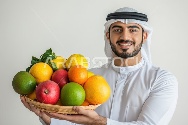 الحفاظ على الصحة وتقليل خطر الاصابة بالأمراض، بورتريه مقرب لشاب عربي خليجي سعودي يرتدي الزي التقليدي والشماغ يمسك بيديه طبق من الفاكهه، محاصيل زراعية سعودية ذات جودة عالية، تجنب المأكولات الضارة، توفير العناصر الغذائية الضرورية للجسم، خلفية رماديه