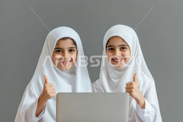 متابعة حصص الاونلاين عن طريق جهاز الحاسوب، بورتريه لطالبتان عربيتان خليجيتان سعوديتان محجبتان يستخدمان الحاسب المحمول وينظران إلى الكاميرا بإيماءات السعادة والسرور يرفعن اصبع الابهام الى الاعلى، التعلم عن بعد باستخدام التكنولوجيا الحديثة، خلفية بيضاء