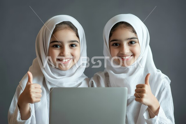 متابعة حصص الاونلاين عن طريق جهاز الحاسوب، بورتريه لطالبتان عربيتان خليجيتان سعوديتان محجبتان يستخدمان الحاسب المحمول وينظران إلى الكاميرا بإيماءات السعادة والسرور يرفعن اصبع الابهام الى الاعلى، التعلم عن بعد باستخدام التكنولوجيا الحديثة، خلفية بيضاء