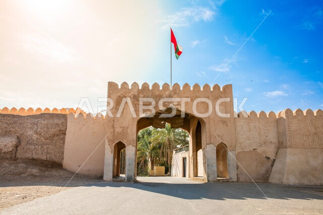 Structural engineering designs of ancient forts and castles, mud gates built in the old style, a famous tourist destination to learn about the country's history and civilization, interest in planting palm trees, the flag of the Sultanate of Oman fluttering above the castle gate