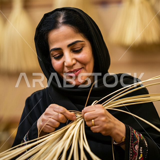 التراث والأصالة في السعودية، استخدام أوراق النخيل المجففة في صناعة السلل، منتجات محلية وطنية، مهن نسائية شعبية تقليدية، إمرأة عربية خليجية سعودية ترتدي الحجاب والعباءة تقوم بجدل سعائف الخوص، حرف يدوية قديمة تاريخية