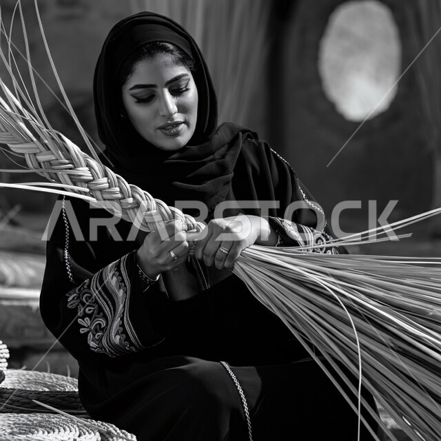 استخدام أوراق النخيل المجففة في صناعة السلل، التراث والأصالة في السعودية، منتجات محلية وطنية، مهن نسائية شعبية تقليدية، إمرأة عربية خليجية سعودية ترتدي الحجاب والعباءة تقوم بجدل سعائف الخوص، حرف يدوية قديمة تاريخية
