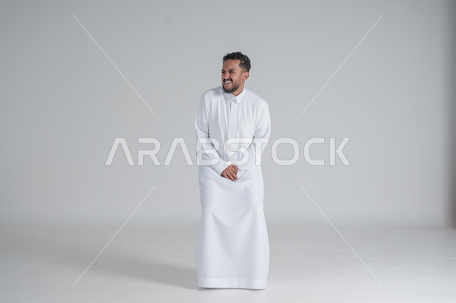 ايماءات الالم والوجع، الحاجه للذهاب الى دورة المياه، بورتريه لشاب عربي سعودي خليجي يرتدي الثوب التقليدي يضع يديه اسفل بطنه بشدة ينظر الى شيء ما, صورة الجسم بالطول الكامل، خلفية بيضاء