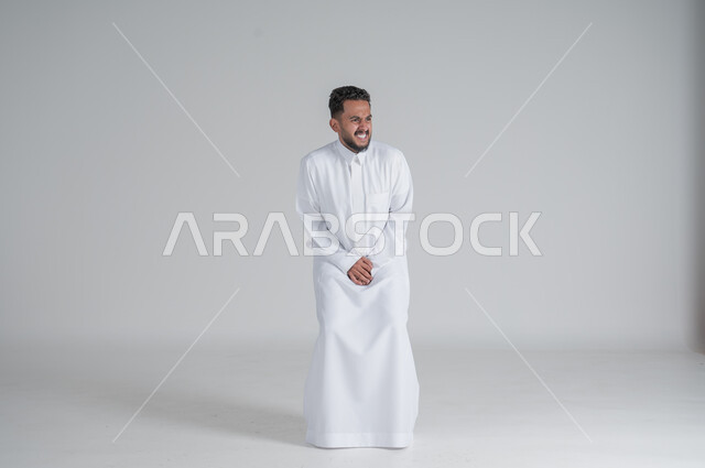ايماءات الالم والوجع، الحاجه للذهاب الى دورة المياه، بورتريه لشاب عربي سعودي خليجي يرتدي الثوب التقليدي يضع يديه اسفل بطنه بشدة ينظر الى شيء ما, صورة الجسم بالطول الكامل، خلفية بيضاء