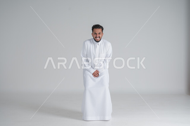 Gestures of pain and suffering, need to go to the bathroom, portrait of a young Saudi Arabian Gulf man wearing traditional thobe, putting his hands under his stomach tightly, looking at the camera, full body photo, white background