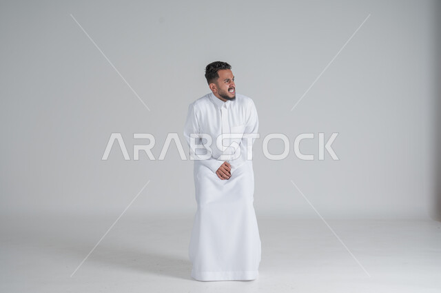 ايماءات الالم والوجع، الحاجه للذهاب الى دورة المياه، بورتريه لشاب عربي سعودي خليجي يرتدي الثوب التقليدي يضع يديه اسفل بطنه بشدة ينظر الى شيء ما, صورة الجسم بالطول الكامل، خلفية بيضاء