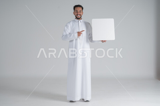 إستخدام لوحات توضيحية ووسائل تعليمية، إعلانات وعروضات تسويقية، بورتريه لشاب عربي سعودي خليجي يرتدي الثوب التقليدي يحمل بيده لوحه بيضاء مربعة ويشير بأصبع السبابة اليها بإيماءات السرور، موك اب مربع أبيض، لوح إعلاني فارغ، صورة الجسم بالطول الكامل, خلفية بيضاء