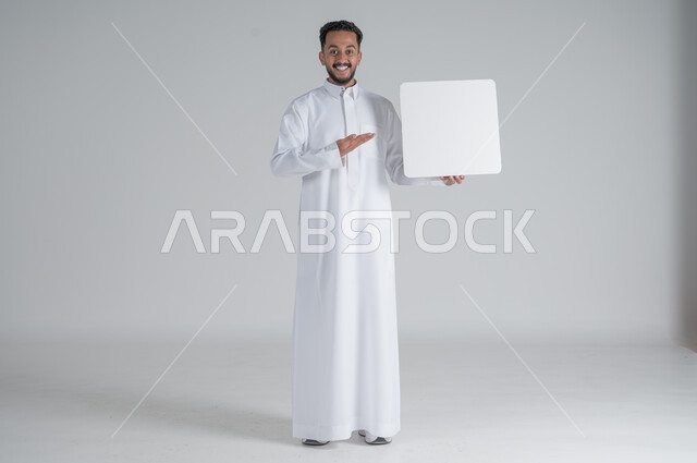 إستخدام لوحات توضيحية ووسائل تعليمية، إعلانات وعروضات تسويقية، بورتريه لشاب عربي سعودي خليجي يرتدي الثوب التقليدي يحمل بيده لوحه بيضاء مربعة ويشير بيده اليها بإيماءات السرور، موك اب مربع أبيض، لوح إعلاني فارغ، صورة الجسم بالطول الكامل, خلفية بيضاء