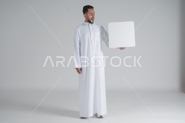 Using illustrations and educational tools, advertisements and marketing offers, portrait of a young Saudi Arabian Gulf man wearing traditional dress holding a square white board with gestures of surprise and shock, white square mockup, blank billboard, full-length body image, white background