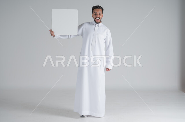 Using illustrations and educational tools, advertisements and marketing offers, portrait of a young Saudi Arabian Gulf man wearing traditional dress holding a square white board with gestures of surprise and shock, white square mockup, blank billboard, full-length body image, white background