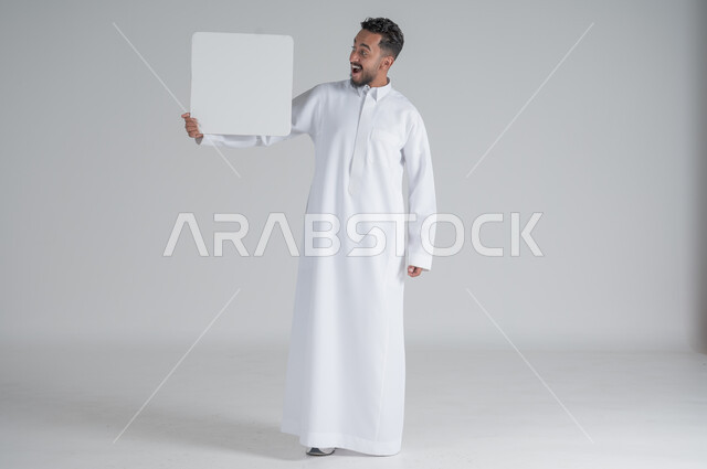 إستخدام لوحات توضيحية ووسائل تعليمية، إعلانات وعروضات تسويقية، بورتريه لشاب عربي سعودي خليجي يرتدي الثوب التقليدي يحمل بيده لوحه بيضاء مربعة بإيماءات المفاجئة والصدمة، موك اب مربع أبيض، لوح إعلاني فارغ، صورة الجسم بالطول الكامل, خلفية بيضاء