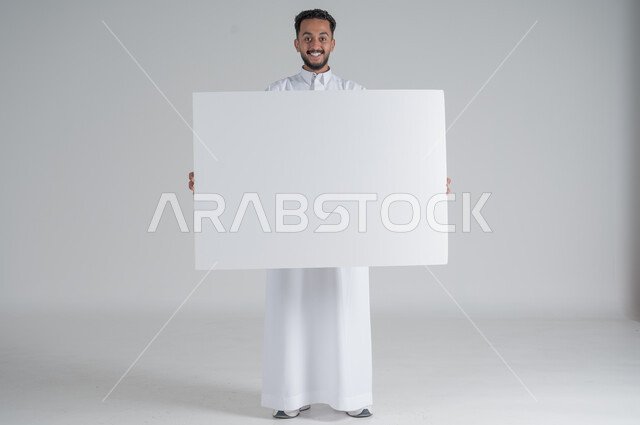 إستخدام لوحات توضيحية ووسائل تعليمية، إعلانات وعروضات تسويقية، بورتريه لشاب عربي سعودي خليجي يرتدي الثوب التقليدي يحمل بيده لوحه بيضاء مستطيلة بإيماءات السرور، موك اب مستطيل أبيض، لوح إعلاني فارغ، صورة الجسم بالطول الكامل, خلفية بيضاء
