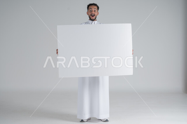 إستخدام لوحات توضيحية ووسائل تعليمية، إعلانات وعروضات تسويقية، بورتريه لشاب عربي سعودي خليجي يرتدي الثوب التقليدي يحمل بيده لوحه بيضاء مستطيلة بإيماءات المفاجئة و الصدمة، موك اب مستطيل أبيض، لوح إعلاني فارغ، صورة الجسم بالطول الكامل, خلفية بيضاء
