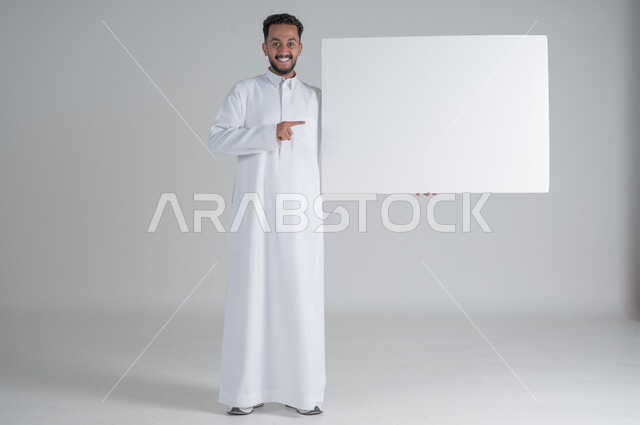 إعلانات وعروضات تسويقية، بورتريه لشاب عربي سعودي خليجي يرتدي الثوب التقليدي يحمل بيده لوحه بيضاء مستطيلة ويشير اليها بأصبغ السبابة بإيماءات السرور، إستخدام لوحات توضيحية ووسائل تعليمية، موك اب مستطيل أبيض، لوح إعلاني فارغ، صورة الجسم بالطول الكامل, خلفية بيضاء