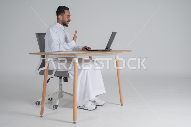 Using modern and advanced technologies in office work, responding to inquiries and questions and customer service, Saudi administrative professions and jobs, portrait of a young Saudi Arabian Gulf man wearing traditional dress sitting on a chair in front of a laptop, gestures of happiness and pleasure, white background