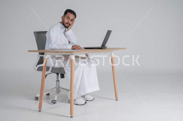 ايماءات الملل والضجر، استخدام التقنيات الحديثة والمتطورة بالأعمال المكتبية، الرد على الاستعلامات والاستفسارات وخدمة العملاء، مهن ووظائف إدارية سعودية، بورتريه لشاب عربي سعودي خليجي يرتدي الثوب التقليدي يجلس على الكرسي أمام الحاسوب المحمول يضع يده على خده،  خلفية بيضاء