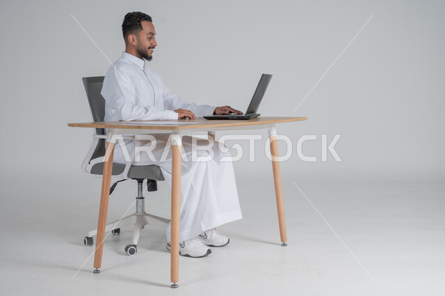 استخدام التقنيات الحديثة والمتطورة بالأعمال المكتبية، الرد على الاستعلامات والاستفسارات وخدمة العملاء، مهن ووظائف إدارية سعودية، بورتريه لشاب عربي سعودي خليجي يرتدي الثوب التقليدي يجلس على الكرسي أمام الحاسوب المحمول ، ايماءات السعادة والسرور، خلفية بيضاء