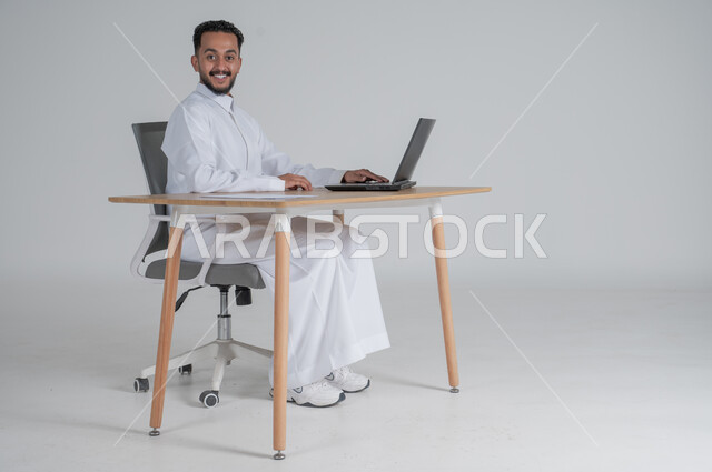 استخدام التقنيات الحديثة والمتطورة، بورتريه لشاب عربي سعودي خليجي يرتدي الثوب التقليدي يضع سماعة الهيدفون و يعمل عن بعد باستخدام الحاسوب المحمول ينظر الى الكاميرا بإيماءات السرور، بيئة العمل السعودي، خلفية بيضاء