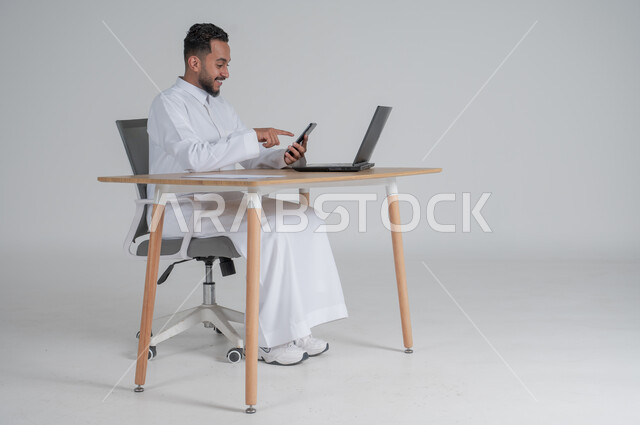 استخدام التقنيات الحديثة والمتطورة، بورتريه لشاب عربي سعودي خليجي يرتدي الثوب التقليدي يجلس على الكرسي أمام الحاسوب المحمول مبتسم يحمل بيده الجوال ويشير اليه، ايماءات السعادة والسرور، الإجابة على الاستعلامات والاستفسارات، خلفية بيضاء
