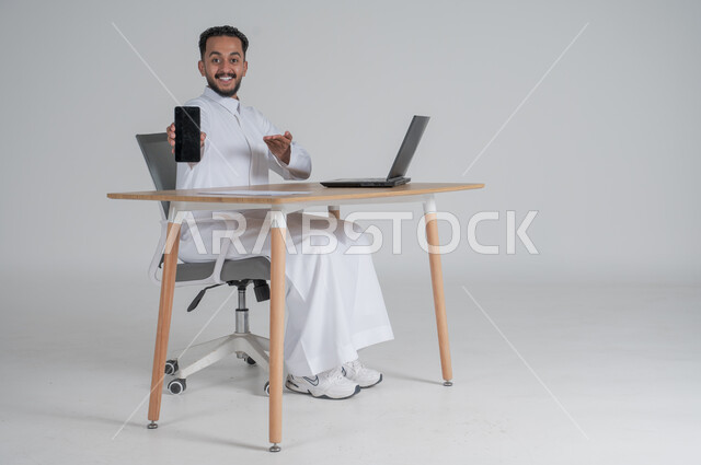 استخدام التقنيات الحديثة والمتطورة، بورتريه لشاب عربي سعودي خليجي يرتدي الثوب التقليدي يجلس على الكرسي أمام الحاسوب المحمول مبتسم يحمل بيده الجوال بشاشة سوداء فارغه ويشير اليه بيديه، النظر الى الكاميرا بايماءات السعادة والسرور، خلفية بيضاء