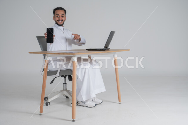 استخدام التقنيات الحديثة والمتطورة، بورتريه لشاب عربي سعودي خليجي يرتدي الثوب التقليدي يجلس على الكرسي أمام الحاسوب المحمول مبتسم يحمل بيده الجوال بشاشة سوداء فارغه ويشير اليه بأصبع السبابة، النظر الى الكاميرا بايماءات السعادة والسرور، خلفية بيضاء