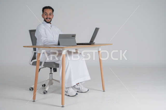 Making a video call via tablet, using modern and advanced technologies, portrait of a young Saudi Arabian Gulf man wearing traditional thobe, wearing headphones and working remotely using a laptop, pointing with his index fingers, Saudi work environment, white background