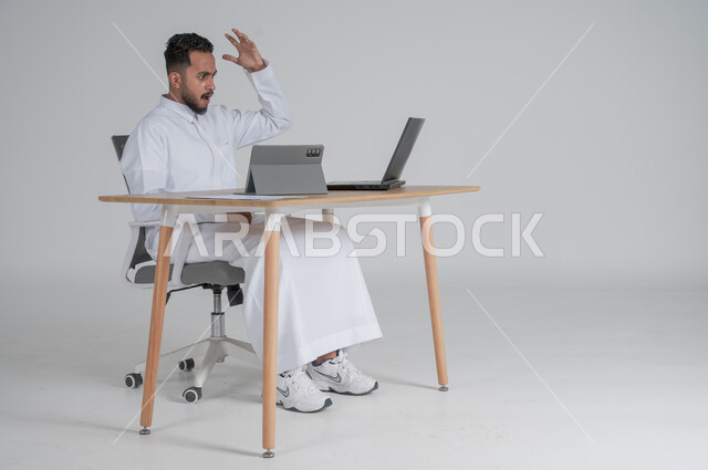استخدام التقنيات الحديثة والمتطورة، إجراء مكالمة فيديو عبر التابليت اللوحي، بورتريه لشاب عربي سعودي خليجي يرتدي الثوب التقليدي يضع سماعة الهيدفون و يعمل عن بعد باستخدام الحاسوب المحمول، ايماءات الغضب و العصبية، بيئة العمل السعودي، خلفية بيضاء