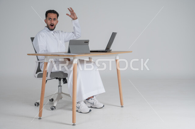 استخدام التقنيات الحديثة والمتطورة، إجراء مكالمة فيديو عبر التابليت اللوحي، بورتريه لشاب عربي سعودي خليجي يرتدي الثوب التقليدي يضع سماعة الهيدفون و يعمل عن بعد باستخدام الحاسوب المحمول، ايماءات الغضب و العصبية، بيئة العمل السعودي، خلفية بيضاء