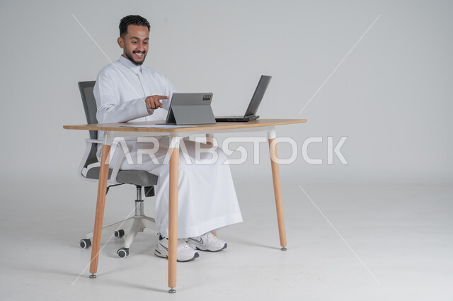 Making a video call via tablet, using modern and advanced technologies, portrait of a young Saudi Arabian Gulf man wearing traditional thobe, wearing headphones and working remotely using a laptop, pointing with his index fingers, Saudi work environment, white background
