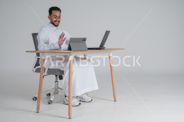 إجراء مكالمة فيديو عبر التابليت اللوحي، استخدام التقنيات الحديثة والمتطورة، بورتريه لشاب عربي سعودي خليجي يرتدي الثوب التقليدي يضع سماعة الهيدفون و يعمل عن بعد باستخدام الحاسوب المحمول يشير بأصبعيه السبابة، بيئة العمل السعودي، خلفية بيضاء