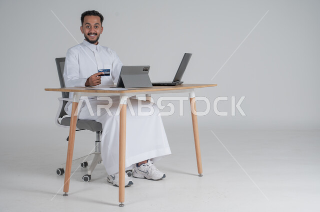 إمكانية الشراء والدفع من خلال البرامج والتطبيقات الالكترونية، استخدام اجهزة تقنية حديثة للدفع عن بعد عبر الانترنت، بورتريه لشاب عربي سعودي خليجي يرتدي الثوب التقليدي يضع سماعة الهيدفون يجلس على كرسي مريح أمام الحاسوب المحمول والتابليت اللوحي يحمل بطاقة الائتمان البنكية للتسوق الالكتروني، خلفية بيضاء