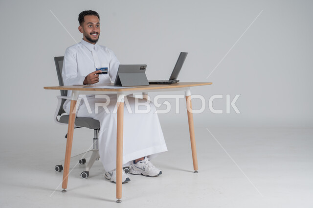 The possibility of purchasing and paying through electronic programs and applications, using modern technical devices for remote payment via the Internet, a portrait of a young Saudi Arabian Gulf man wearing traditional clothing, wearing headphones, sitting on a comfortable chair in front of a laptop and tablet holding a bank credit card for electronic shopping, white background