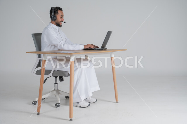 الرد على الاستعلامات والاستفسارات وخدمة العملاء، استخدام التقنيات الحديثة والمتطورة، بورتريه لشاب عربي سعودي خليجي يرتدي الثوب التقليدي يضع سماعة الهيدفون و يعمل عن بعد باستخدام الحاسوب المحمول، بيئة العمل السعودي، خلفية بيضاء