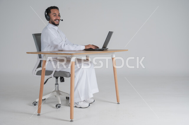 الرد على الاستعلامات والاستفسارات وخدمة العملاء، استخدام التقنيات الحديثة والمتطورة، بورتريه لشاب عربي سعودي خليجي يرتدي الثوب التقليدي يضع سماعة الهيدفون و يعمل عن بعد باستخدام الحاسوب المحمول، بيئة العمل السعودي، خلفية بيضاء