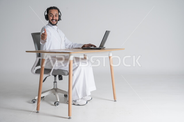 استخدام التقنيات الحديثة والمتطورة، بورتريه لشاب عربي سعودي خليجي يرتدي الثوب التقليدي يضع سماعة الهيدفون يرفع اصبع الابهام بتعابير الإعجاب والرضى و الموافقة، بيئة العمل السعودي، الرد على الاستعلامات والاستفسارات وخدمة العملاء، خلفية بيضاء