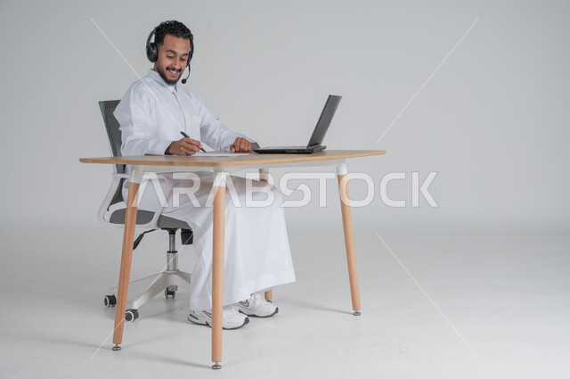 Completing work tasks and taking notes and data, using modern and advanced technologies, portrait of a young Saudi Arabian Gulf man wearing traditional dress, wearing headphones and working remotely, Saudi work environment, responding to inquiries and questions and customer service, white background