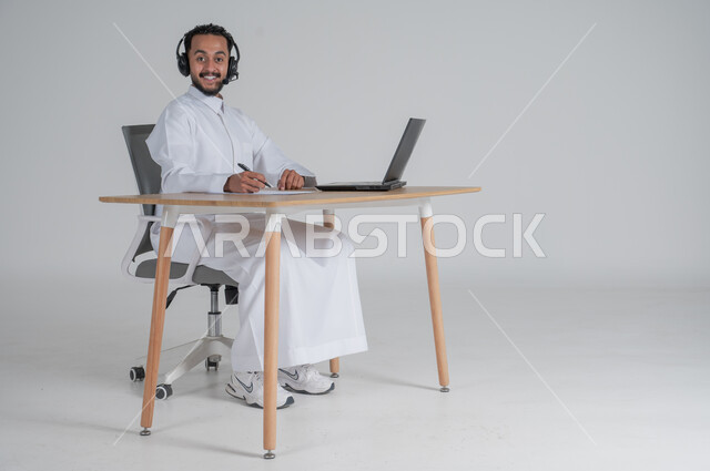 Completing work tasks and taking notes and data, using modern and advanced technologies, portrait of a young Saudi Arabian Gulf man wearing traditional dress, wearing headphones and working remotely, Saudi work environment, responding to inquiries and questions and customer service, white background