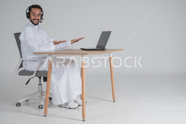 الرد على الاستعلامات والاستفسارات وخدمة العملاء، استخدام التقنيات الحديثة والمتطورة، بورتريه لشاب عربي سعودي خليجي يرتدي الثوب التقليدي يضع سماعة الهيدفون و يعمل عن بعد يشير بأصبعيه السبابة الى الحاسوب المحمول، بيئة العمل السعودي، خلفية بيضاء