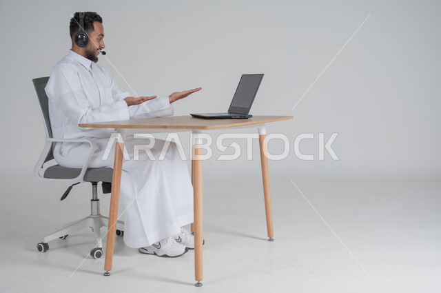 الرد على الاستعلامات والاستفسارات وخدمة العملاء، استخدام التقنيات الحديثة والمتطورة، بورتريه لشاب عربي سعودي خليجي يرتدي الثوب التقليدي يضع سماعة الهيدفون و يعمل عن بعد يشير بأصبعيه السبابة الى الحاسوب المحمول، بيئة العمل السعودي، خلفية بيضاء
