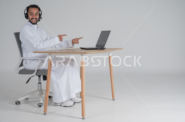الرد على الاستعلامات والاستفسارات وخدمة العملاء، استخدام التقنيات الحديثة والمتطورة، بورتريه لشاب عربي سعودي خليجي يرتدي الثوب التقليدي يضع سماعة الهيدفون و يعمل عن بعد يشير بأصبعيه السبابة الى الحاسوب المحمول، بيئة العمل السعودي، خلفية بيضاء