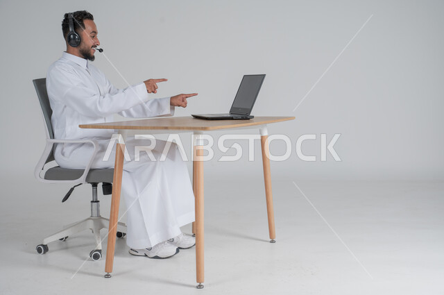 إجراء مكالمة فيديو عبر جهاز الكمبيوتر، استخدام التقنيات الحديثة والمتطورة، بورتريه لشاب عربي سعودي خليجي يرتدي الثوب التقليدي يضع سماعة الهيدفون و يعمل عن بعد باستخدام الحاسوب المحمول يشير بأصبعيه السبابة، بيئة العمل السعودي، خلفية بيضاء