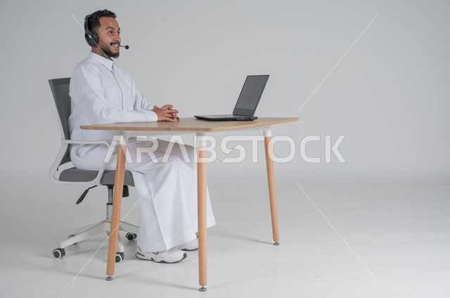 الرد على الاستعلامات والاستفسارات وخدمة العملاء، استخدام التقنيات الحديثة والمتطورة، بورتريه لشاب عربي سعودي خليجي يرتدي الثوب التقليدي يضع سماعة الهيدفون و يعمل عن بعد باستخدام الحاسوب المحمول، بيئة العمل السعودي، خلفية بيضاء