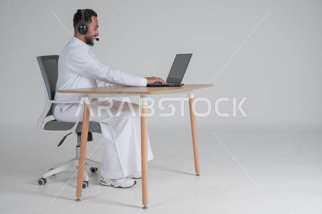 استخدام التقنيات الحديثة والمتطورة، بورتريه لشاب عربي سعودي خليجي يرتدي الثوب التقليدي يضع سماعة الهيدفون و يعمل عن بعد باستخدام الحاسوب المحمول، بيئة العمل السعودي، خلفية بيضاء