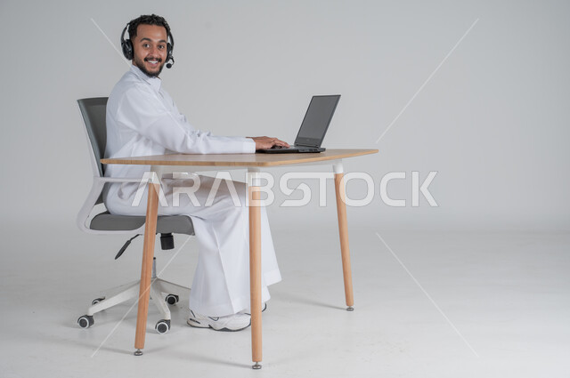 Using modern and advanced technologies, portrait of a young Saudi Arabian Gulf man wearing traditional thobe, wearing headphones and working remotely using a laptop, looking at the camera with gestures of pleasure, Saudi work environment, white background