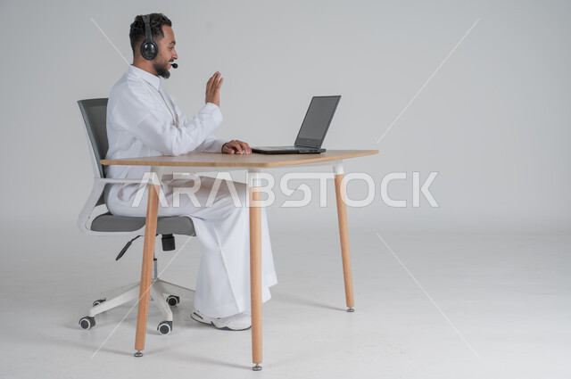 استخدام التقنيات الحديثة والمتطورة، إجراء مكالمة فيديو عبر جهاز الكمبيوتر، بورتريه لشاب عربي سعودي خليجي يرتدي الثوب التقليدي يضع سماعة الهيدفون و يعمل عن بعد باستخدام الحاسوب المحمول، بيئة العمل السعودي، خلفية بيضاء