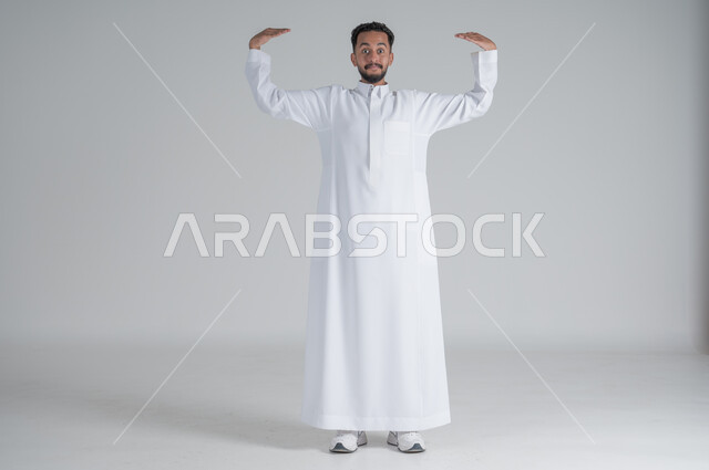 النظر الى الكاميرا بايماءات السعادة والسرور، بورتريه لرجل عربي خليجي سعودي مبتسم يرتدي الثوب التقليدي يرفع يديه للأعلى بايماءات اظهار حجم شيء ما، الجسم بالطول الكامل، خلفية بيضاء