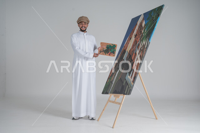 مفهوم الموهبة و الإبداع ,  بورتريه لرسام عربي سعودي خليجي يرتدي الثوب التقليدي ينظر الى الكاميرا بايماءات السعادة والسرور، الشغف في ممارسة الفنون اليدوية و الهوايات المفضلة, استخدام ريشة التلوين والرسم، الجسم بالطول الكامل، خلفية بيضاء