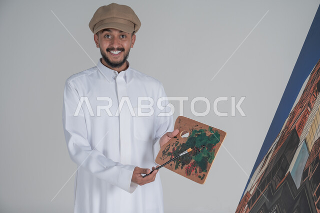 Concept of talent and creativity, close-up portrait of a Saudi Arabian Gulf artist wearing traditional dress looking at the camera with gestures of happiness and pleasure, passion for practicing handicrafts and favorite hobbies, using a paintbrush and drawing, white background